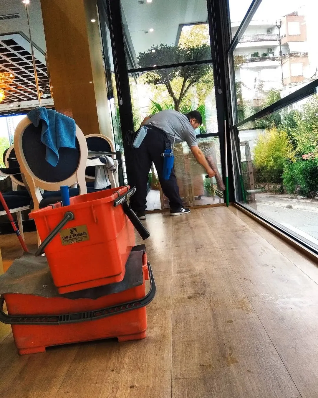 Erjoni nga pas, i përkulur, kalon gomën mbi murin e xhamit të një kafeneje; kova të kuqe në dysheme