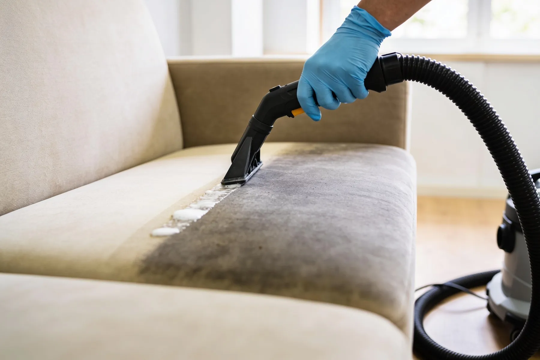 Gloved hand cleaning the seat cushion of a beige fabric sofa with an extraction wand: the cleaned half visibly brighter, the other half still dull